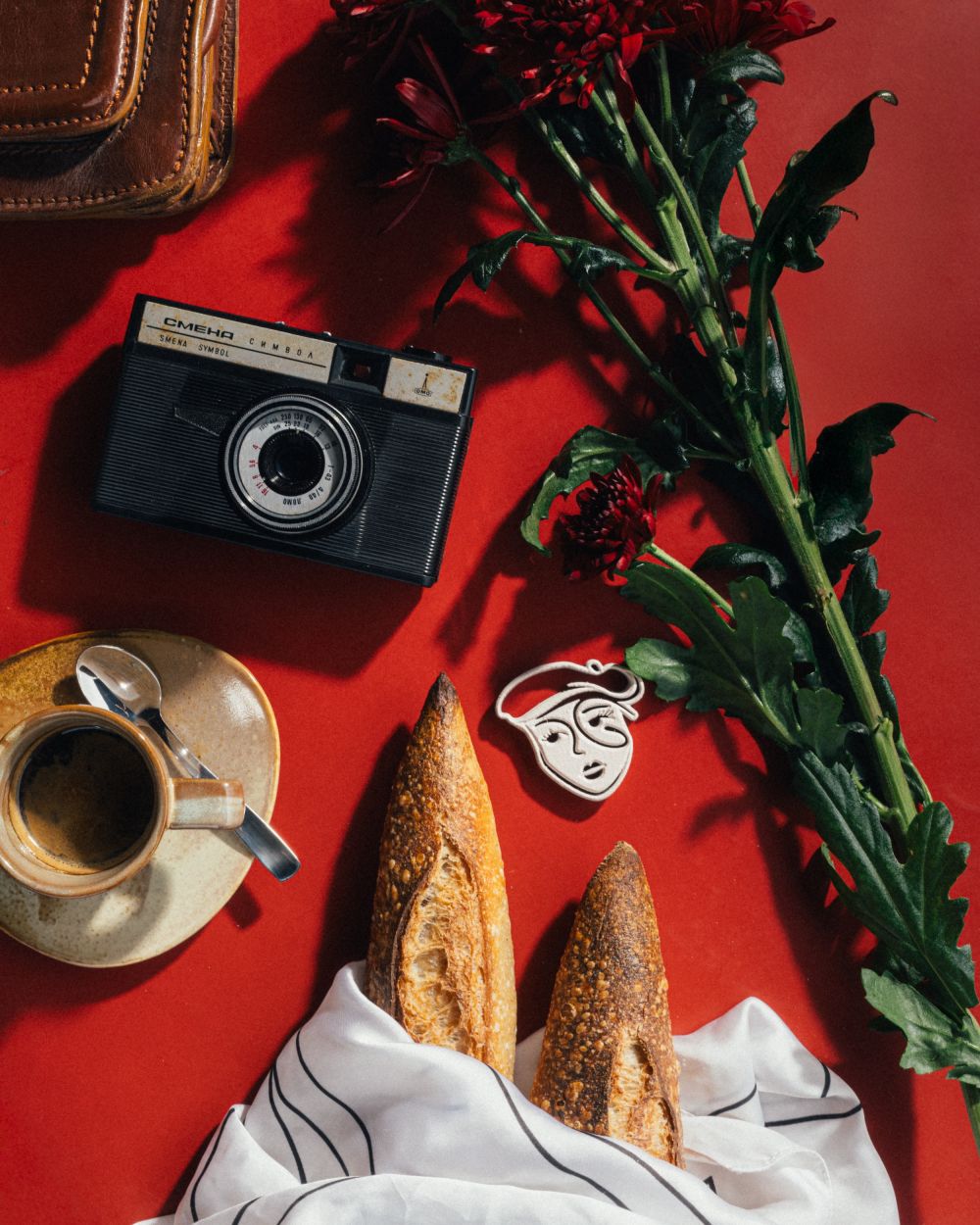 A retro black camera, coffee cup, baguettes wrapped in cloth, and red flowers rest on a table